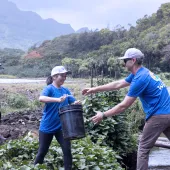 Volunteers in blue shirts pass a bucket across a wooden bridge in a green, mountainous area during a community environmental project.