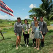 Group of people standing outdoors near a Hawaiian flag and palm tree, with a pond and lush greenery in the background on a sunny day.