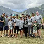 A diverse group of people, including adults and children, pose together outdoors wearing matching gray AES Hawaii t-shirts with lush green mountains and cloudy skies in the background.