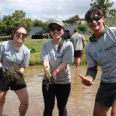 Three people wearing gray shirts and sunglasses smile while planting seedlings in a wet, muddy field during a group outdoor volunteer activity. Other volunteers work in the background under a partly cloudy sky.