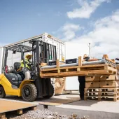 Four construction workers wearing safety vests and helmets operate a forklift to move a large wooden pallet with pipes and equipment at an outdoor construction site on a sunny day.
