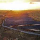Aerial view of a large solar farm in a vast open landscape at sunset, with rows of solar panels capturing sunlight and a road running alongside the facility.