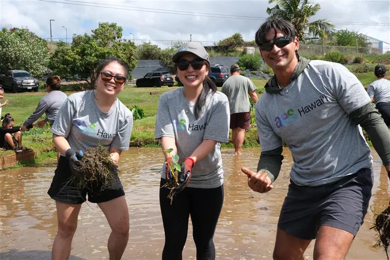 Three people wearing gray shirts and sunglasses smile while planting seedlings in a wet, muddy field during a group outdoor volunteer activity. Other volunteers work in the background under a partly cloudy sky.