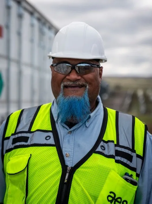 Smiling construction worker with blue beard wearing a white hard hat, safety glasses, and a neon yellow safety vest stands outdoors at a work site on a cloudy day.