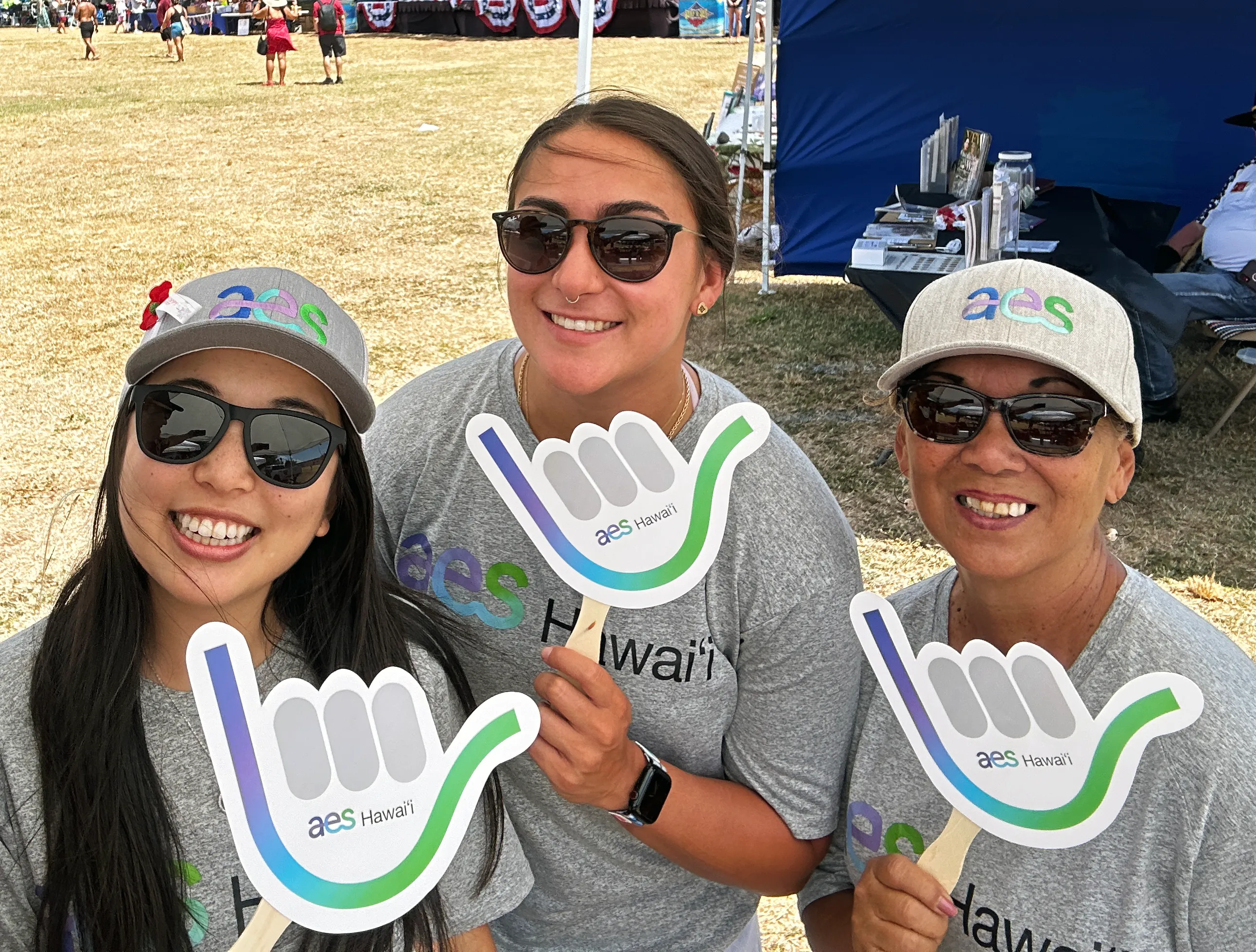 Three people wearing sunglasses and gray shirts with 'aes Hawaii' logos hold shaka sign cutouts at an outdoor event.