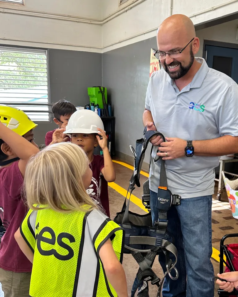 Children in safety gear engage with an adult during a classroom demo, with helmets, bags, and large windows in the background.