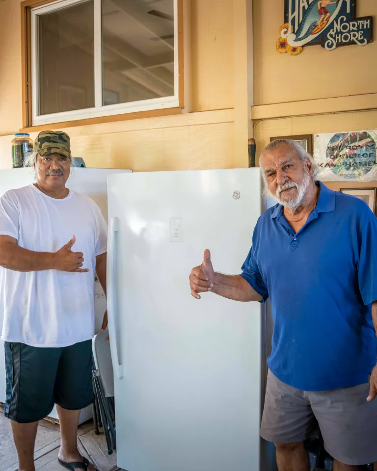 Two people give thumbs-up in front of a white fridge in a covered outdoor space with a dog, shelves, and a TV, near an open door to a green yard.