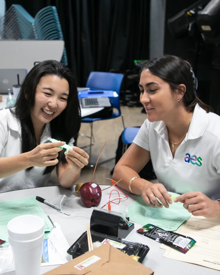Two people at a round table work with electronics, papers, and tools in a workshop or classroom, with supplies and chairs in the background.