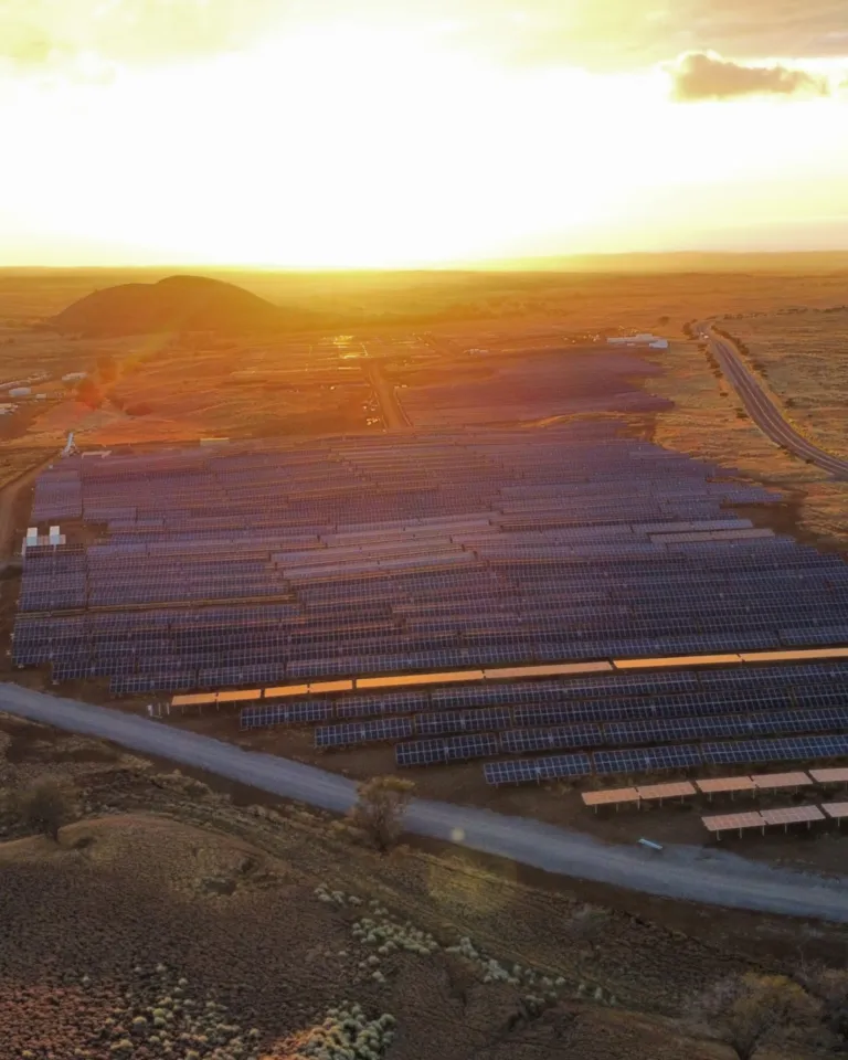 Aerial view of a large solar farm in a vast open landscape at sunset, with rows of solar panels capturing sunlight and a road running alongside the facility.